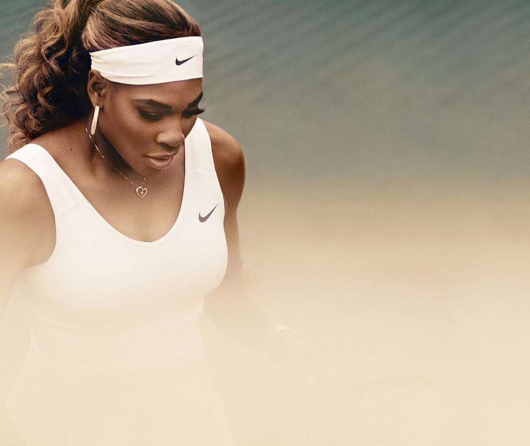 Serena Williams poses wearing white Nike tennis attire, including a headband, tank top, and necklace against a light background.