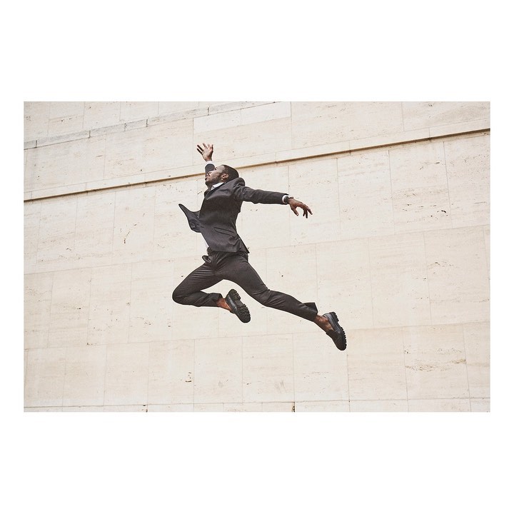 A Black male in business suit leaps mid-air against a minimalist beige wall, showcasing dynamic motion and formal attire in an urban setting.
