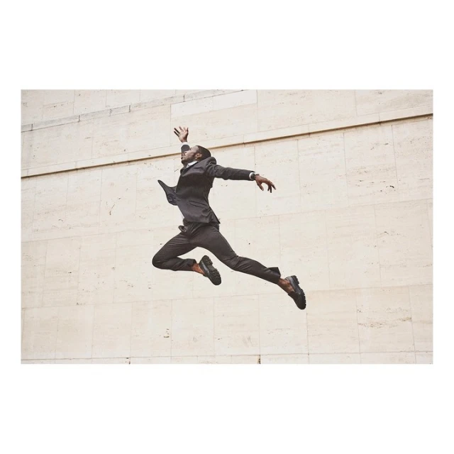 A Black male in business suit leaps mid-air against a minimalist beige wall, showcasing dynamic motion and formal attire in an urban setting.