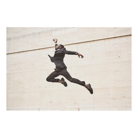A Black male in business suit leaps mid-air against a minimalist beige wall, showcasing dynamic motion and formal attire in an urban setting.