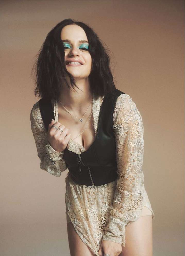 Smiling woman with teal eyeshadow poses indoors in a lace dress and leather vest in a medium shot with a neutral background.