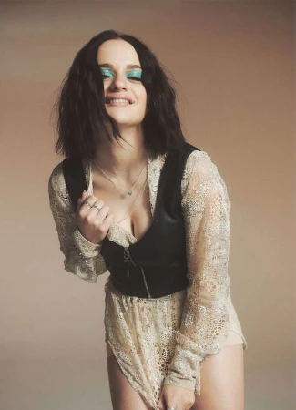 Smiling woman with teal eyeshadow poses indoors in a lace dress and leather vest in a medium shot with a neutral background.