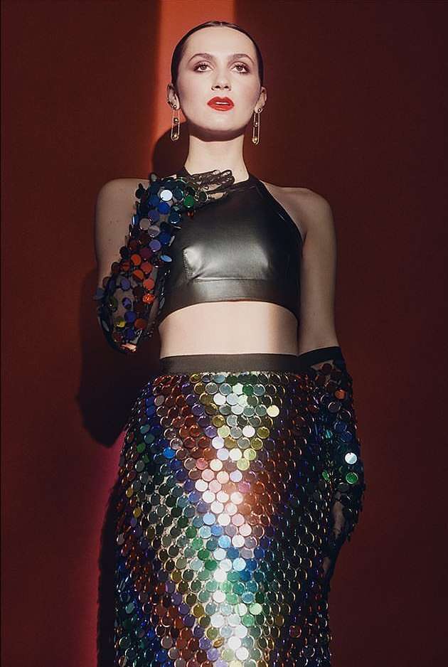 Woman in a black halter top and iridescent skirt poses against a red background, looking up with confidence and style in studio lighting.