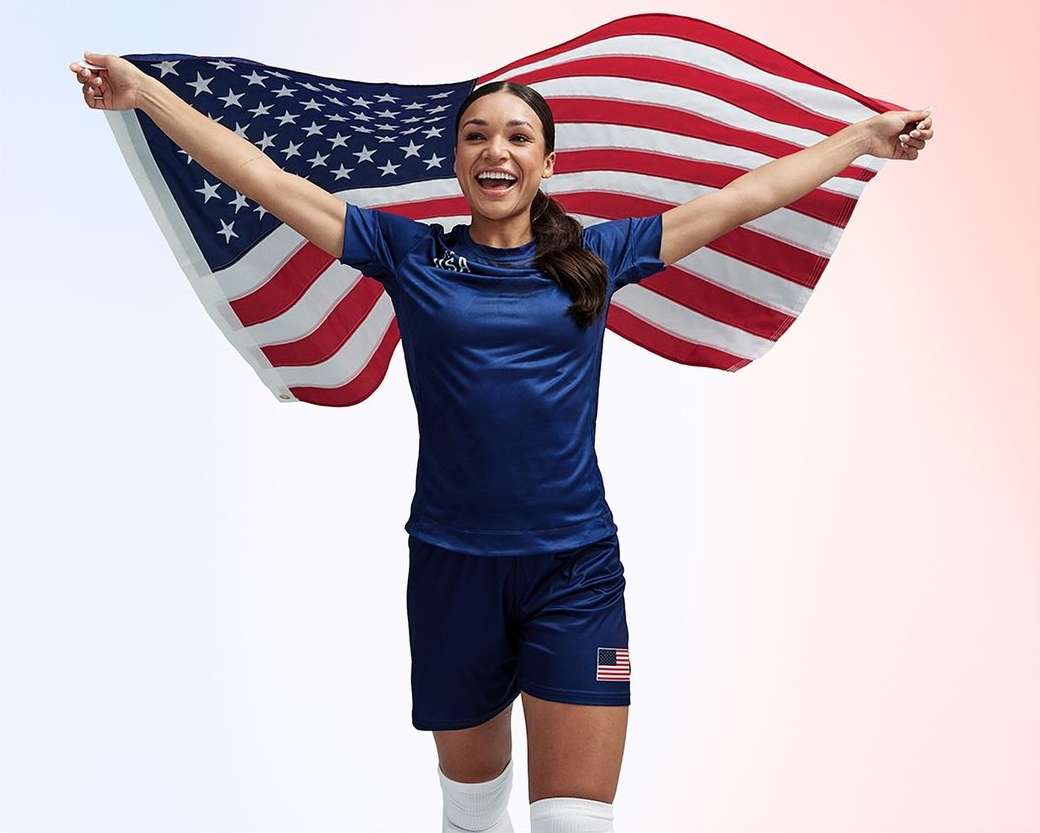 Enthusiastic female athlete stands with arms raised, smiling, while holding an American flag against a gradiated red and white background.