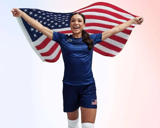 Enthusiastic female athlete stands with arms raised, smiling, while holding an American flag against a gradiated red and white background.