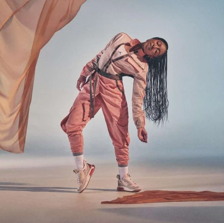 A dancer in pink athletic wear and sneakers arches backward gracefully against an open studio background