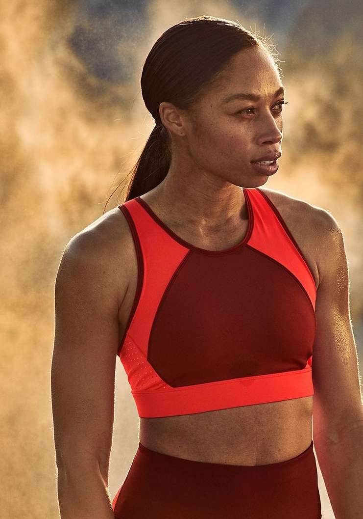 Adult female athlete with dark hair poses in a red sports bra and leggings in a close-up studio shot with a neutral background.
