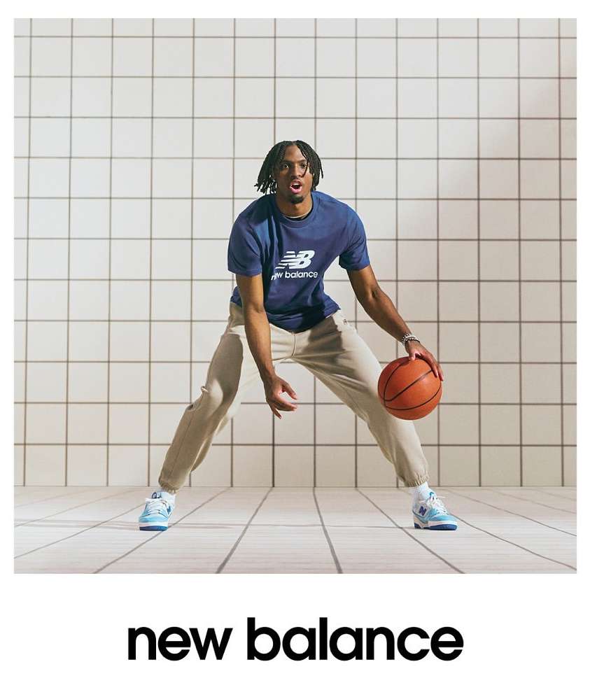 A male athlete dribbles a basketball in a photo studio, wearing a New Balance tee shirt, khakis, and matching NB shoes, promoting the brand.
