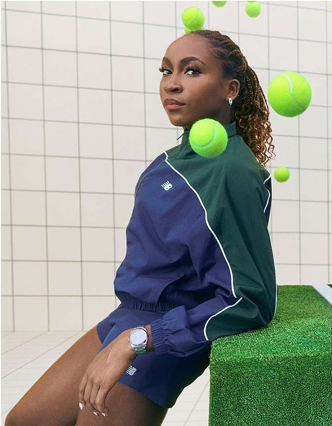 A woman in a navy and green tracksuit poses near floating tennis balls against a white tiled wall, creating a sporty, modern image.