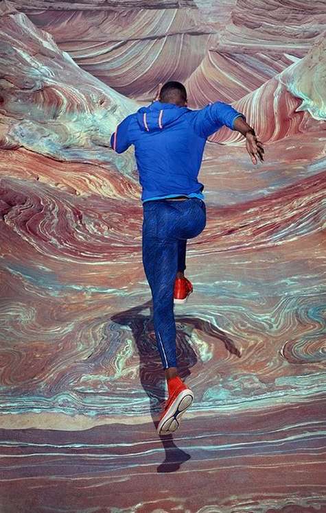 Full-length shot of an adult male in blue activewear jumping in red shoes against a multicolored abstract landscape background with swirling patterns.