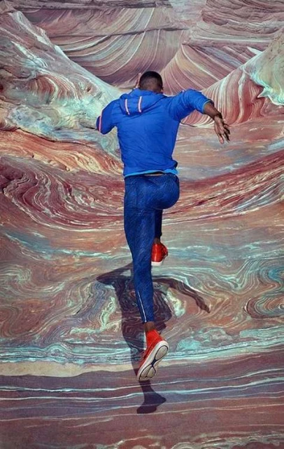 Full-length shot of an adult male in blue activewear jumping in red shoes against a multicolored abstract landscape background with swirling patterns.