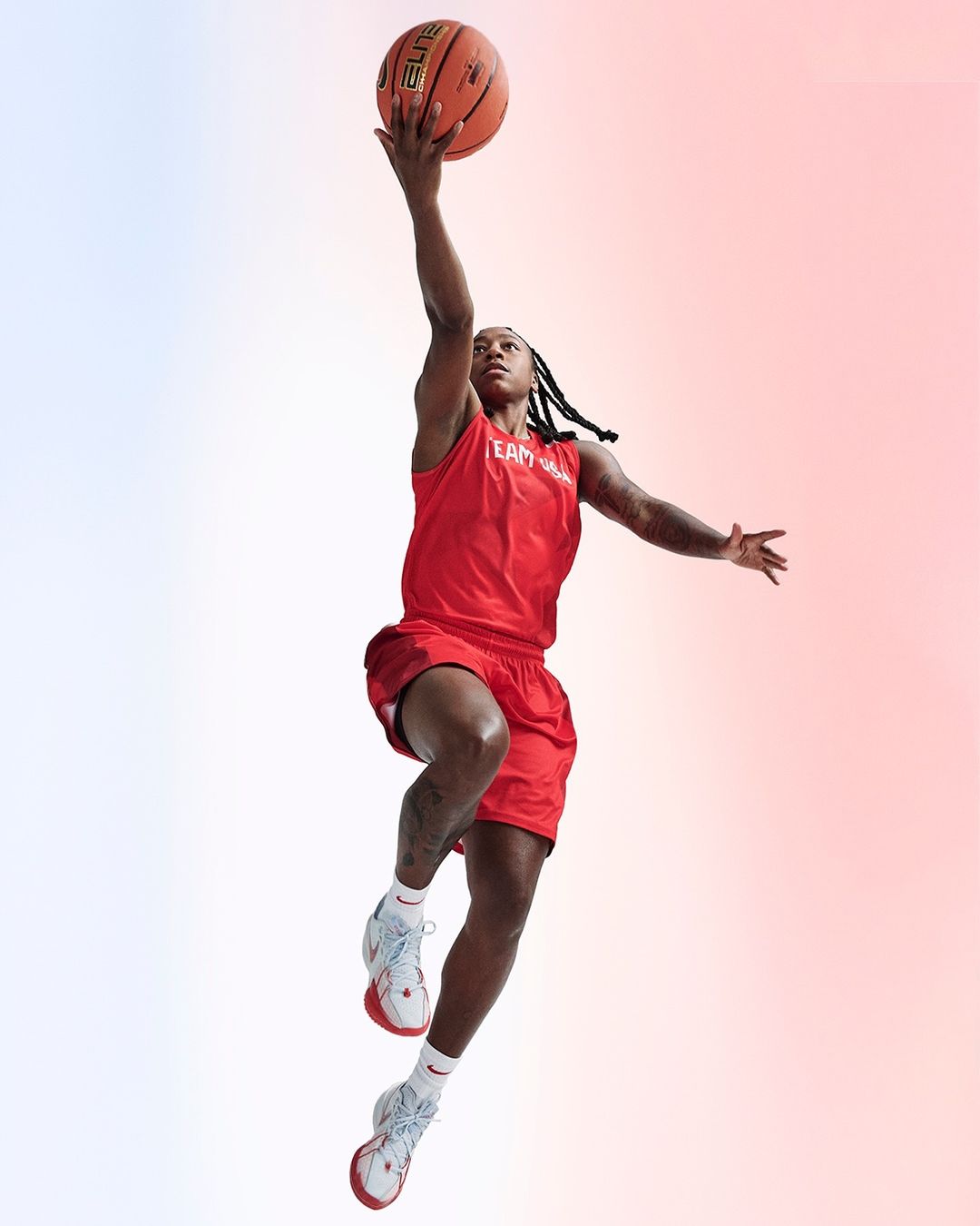 Athletic woman in red sportswear leaps high with a basketball against a gradient background, showcasing her dynamic movement and focus.