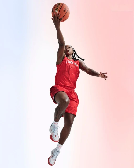 Athletic woman in red sportswear leaps high with a basketball against a gradient background, showcasing her dynamic movement and focus.
