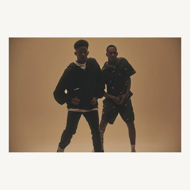 Two black male adults in a casual fashion pose in a photo studio with warm lighting. One is wearing a zip hoodie and black jeans.