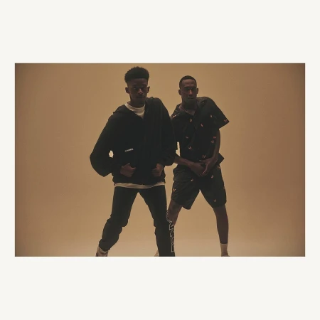 Two black male adults in a casual fashion pose in a photo studio with warm lighting. One is wearing a zip hoodie and black jeans.