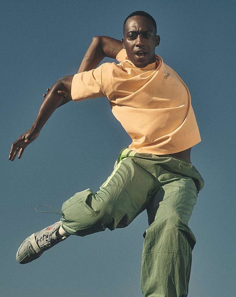 An adult male dancer in mid-air wearing athleisure clothing against a clear blue sky, showcasing a dynamic pose with arms extended for balance.
