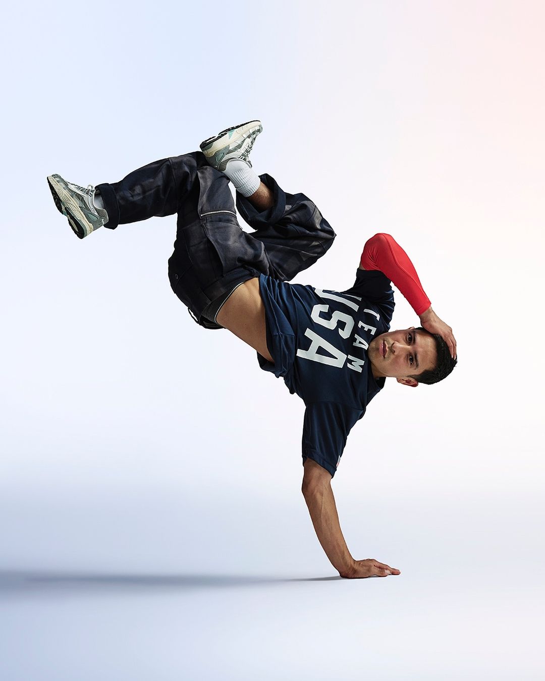 A male breakdancer strikes a dynamic pose in a studio, showcasing his athletic skill, wearing a 'USA Team' t-shirt and sneakers.