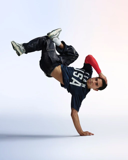 A male breakdancer strikes a dynamic pose in a studio, showcasing his athletic skill, wearing a 'USA Team' t-shirt and sneakers.