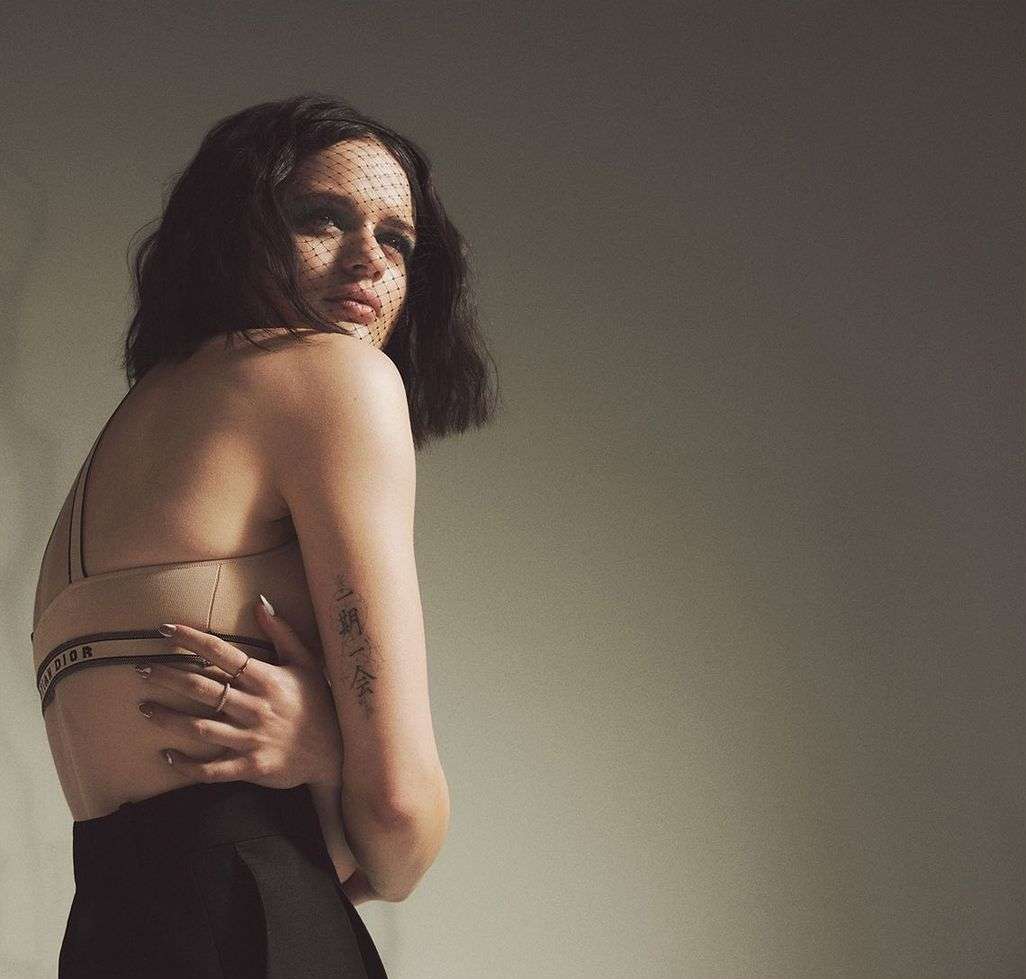 Brunette woman with a veil poses in a tan bra and black skirt, glancing over her shoulder in a studio with soft lighting.