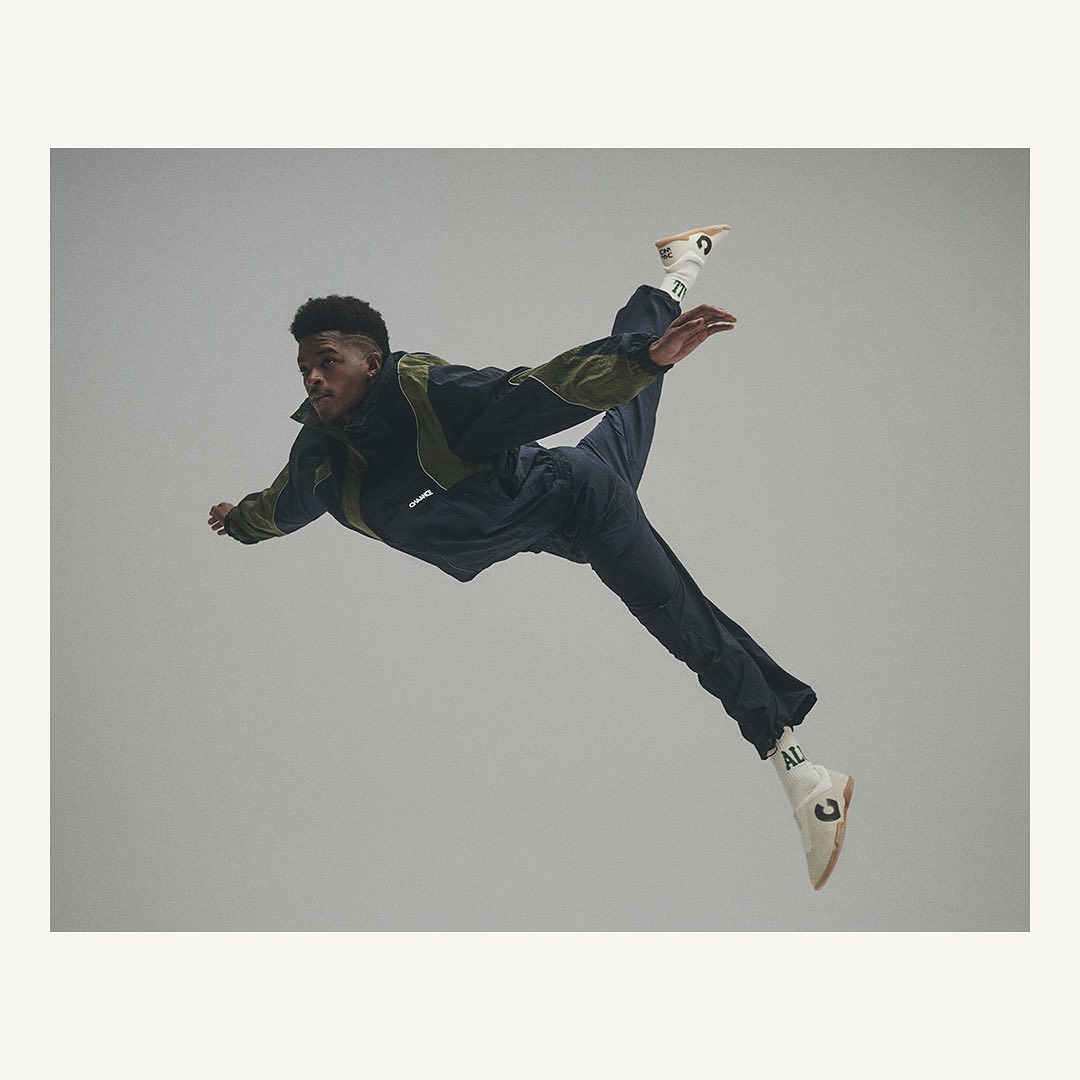 Male dancer in mid-air wearing dark tracksuit and white sneakers, performing a dynamic pose against a neutral grey backdrop.