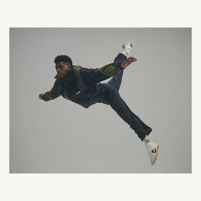 Male dancer in mid-air wearing dark tracksuit and white sneakers, performing a dynamic pose against a neutral grey backdrop.