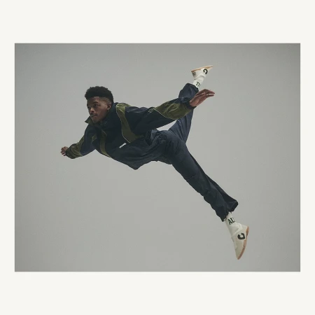 Male dancer in mid-air wearing dark tracksuit and white sneakers, performing a dynamic pose against a neutral grey backdrop.