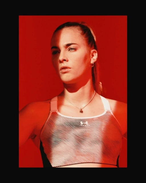 Portrait of a young adult female athlete with blonde hair wearing a sporty top in front of a solid red backdrop.