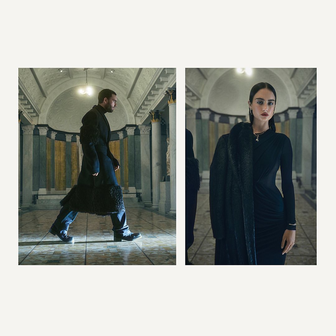 Man in coat walking inside building with woman in black dress and coat, lit by overhead lights, fashion editorial-style photoshoot.