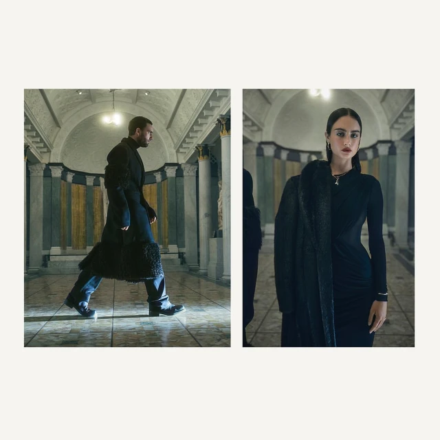 Man in coat walking inside building with woman in black dress and coat, lit by overhead lights, fashion editorial-style photoshoot.