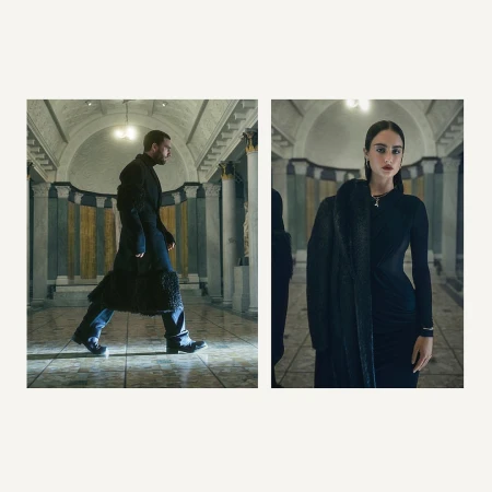 Man in coat walking inside building with woman in black dress and coat, lit by overhead lights, fashion editorial-style photoshoot.