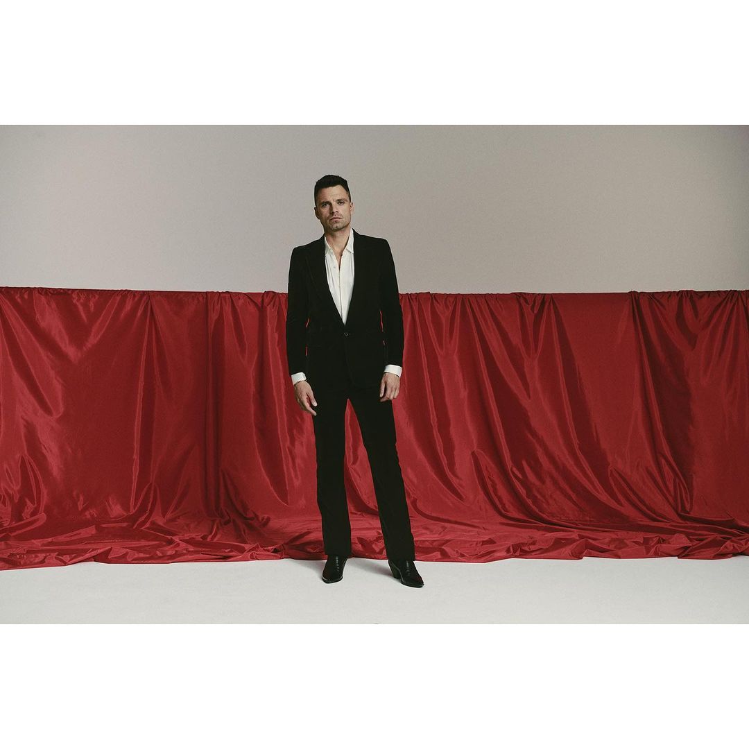A man in a black suit and white shirt stands in front of a red drape against a grey backdrop in a studio shot, looking directly at the camera.