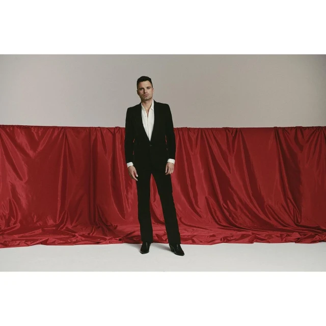 A man in a black suit and white shirt stands in front of a red drape against a grey backdrop in a studio shot, looking directly at the camera.