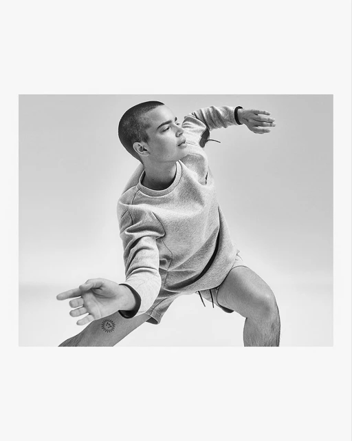 Monochrome portrait of a young adult in grey sweater and shorts, striking a dynamic pose in a studio, with a sun tattoo visible on the arm.