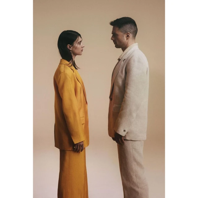 A stylish studio shot showcases a fashionable couple in coordinated suits. The woman in yellow faces the man in beige against a neutral backdrop.
