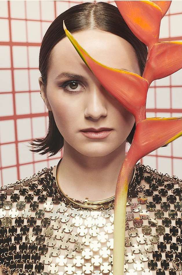 Woman poses with heliconia flower covering eye in studio. She is wearing a reflective top against a tiled coral and white backdrop.