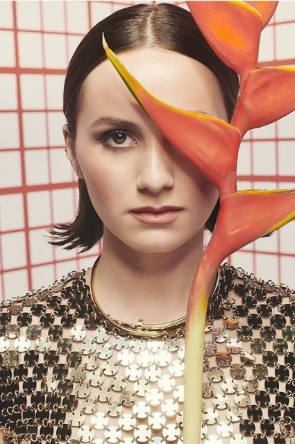 Woman poses with heliconia flower covering eye in studio. She is wearing a reflective top against a tiled coral and white backdrop.