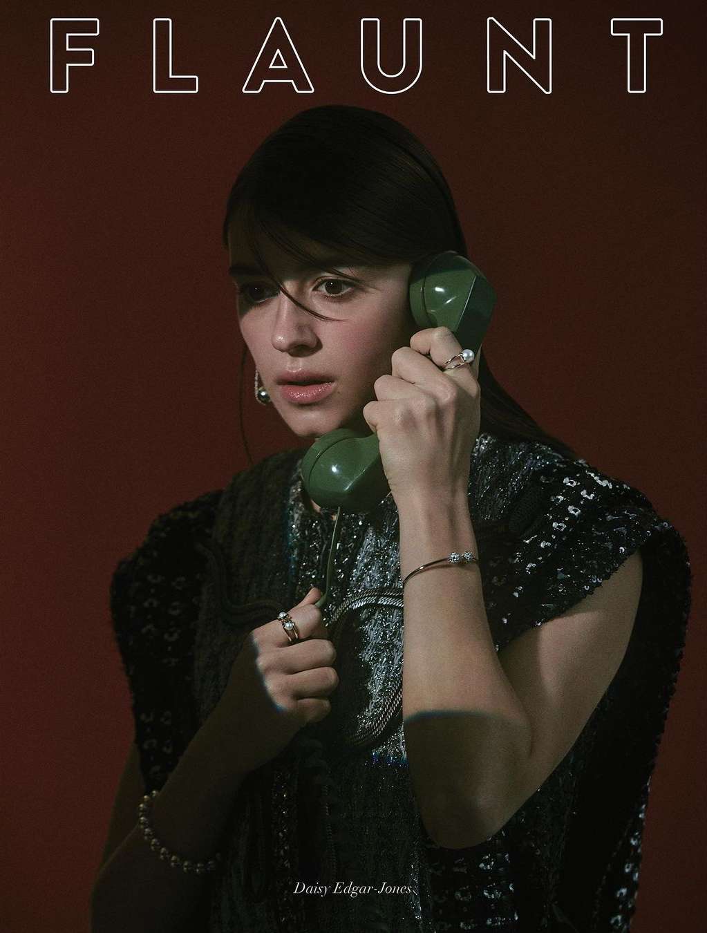 Close-up portrait of Daisy Edgar-Jones holding a vintage green rotary phone, wearing a dark textured sleeveless top with pearl jewelry.