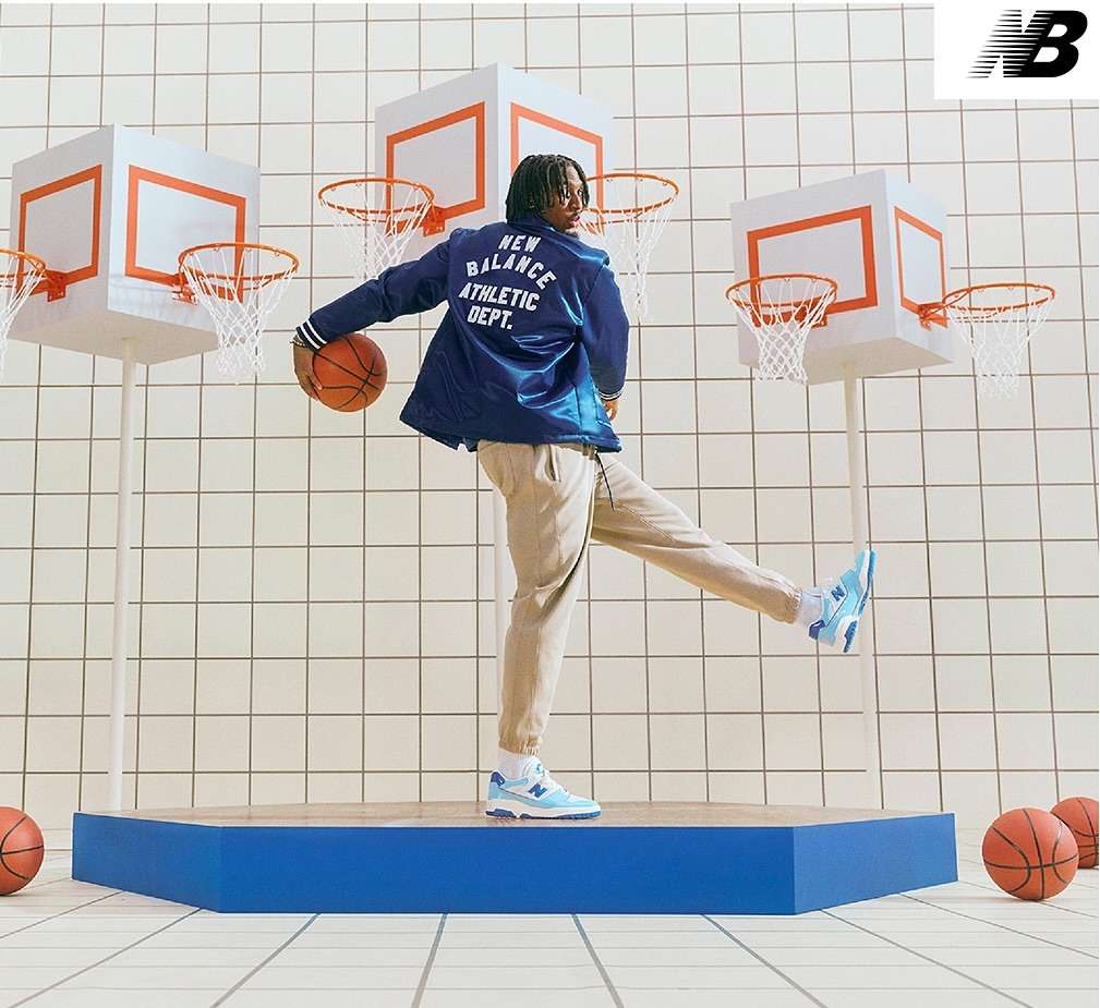 Man in retro blue jacket and sneakers poses with a basketball on a blue platform in front of cube basketball hoops and tiled wall.
