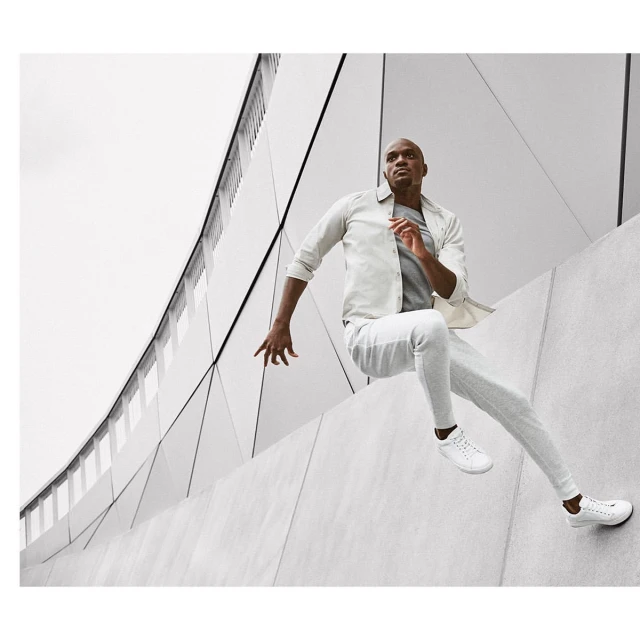 An African-American man leaps over a concrete wall outdoors, wearing a light-colored jacket, pants, sneakers, with a modern building in the background.