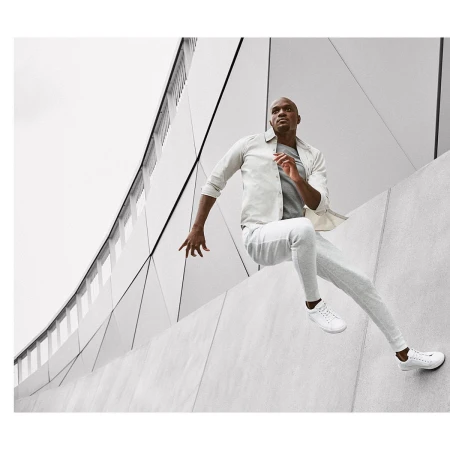 An African-American man leaps over a concrete wall outdoors, wearing a light-colored jacket, pants, sneakers, with a modern building in the background.