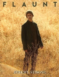 Actor Jeremy Strong wearing a dark jacket and beanie standing in a tall dry meadow during golden hour