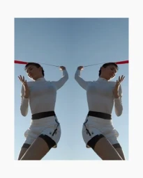 Mirrored symmetry portrait of a woman in a white long-sleeve top and shorts holding a red ribbon against a clear blue sky
