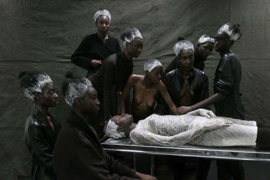 Group of Black models with light painted hair posing around one model on a metal table in front of a solid gray backdrop.