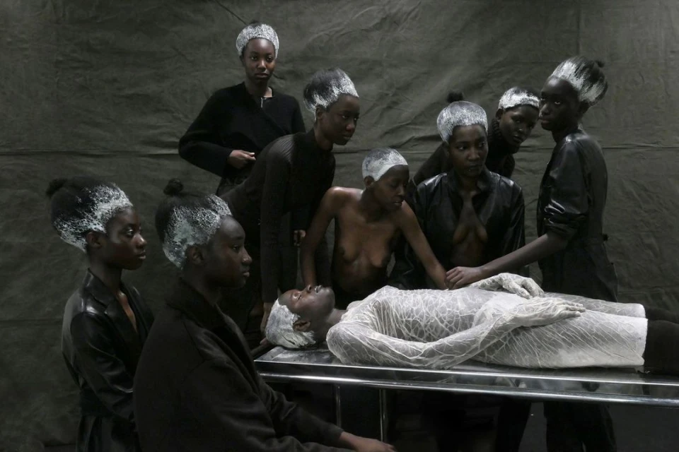 Group of Black models with light painted hair posing around one model on a metal table in front of a solid gray backdrop.