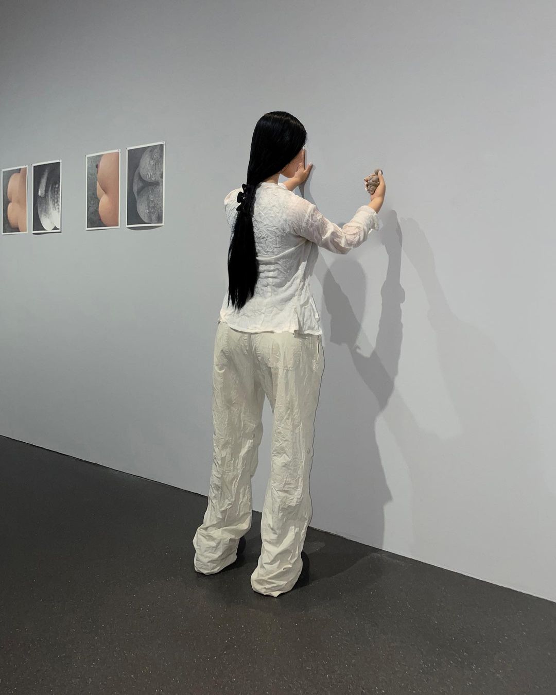 An adult woman with long dark hair is holding an item and looking at art hanging on a gallery wall in a neutral indoor setting.