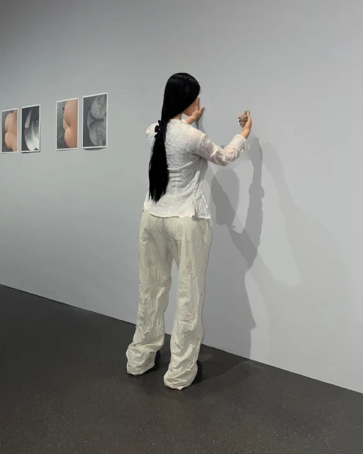 An adult woman with long dark hair is holding an item and looking at art hanging on a gallery wall in a neutral indoor setting.