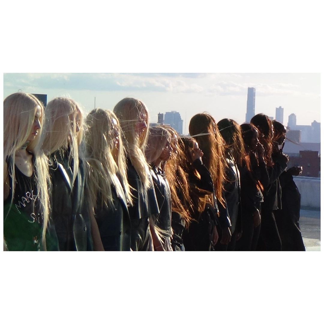 A line of models with long blonde hair standing on a rooftop against a city skyline by Carlota Guerrero.