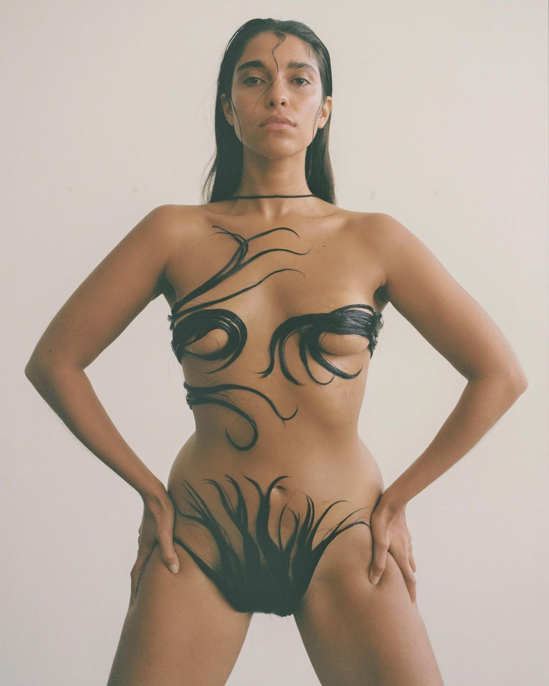 Striking studio shot of a woman with body art: dark, abstract designs on her torso and hips, set against a neutral background.
