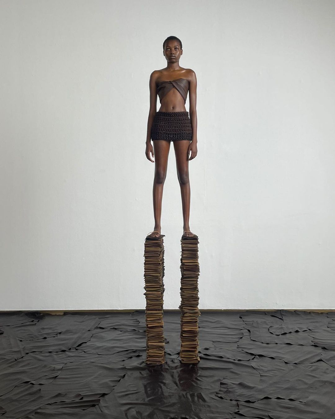 A slender woman in a brown two-piece outfit confidently stands on a stack of books indoors against a white wall, with a dark material on the floor.