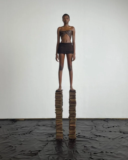 A slender woman in a brown two-piece outfit confidently stands on a stack of books indoors against a white wall, with a dark material on the floor.